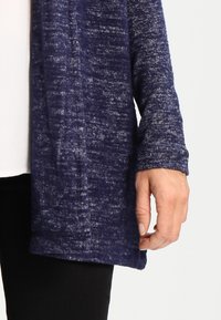 Cardigan blu navy con un punto tessuto, caratterizzato da maniche lunghe e un design aperto davanti. Il materiale sembra morbido e casual.
