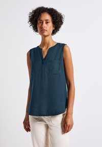 Blusa senza maniche di un verde scuro con scollatura a V, due tasche frontali e spacchi laterali. Realizzata in tessuto morbido con una vestibilità comoda.