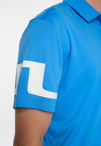 Blå pikétröja med korta ärmar. Har en vit grafik på ärmen. Tillverkad av mjukt, lätt material. Klassisk krage design.