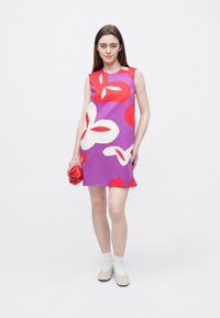 Μωβ αμάνικο φόρεμα με μεγάλα κόκκινα και λευκά λουλουδάτα σχέδια. Το μοντέλο κρατάει ένα κόκκινο αξεσουάρ και φοράει μπεζ παπούτσια με ανοιχτόχρωμες κάλτσες.