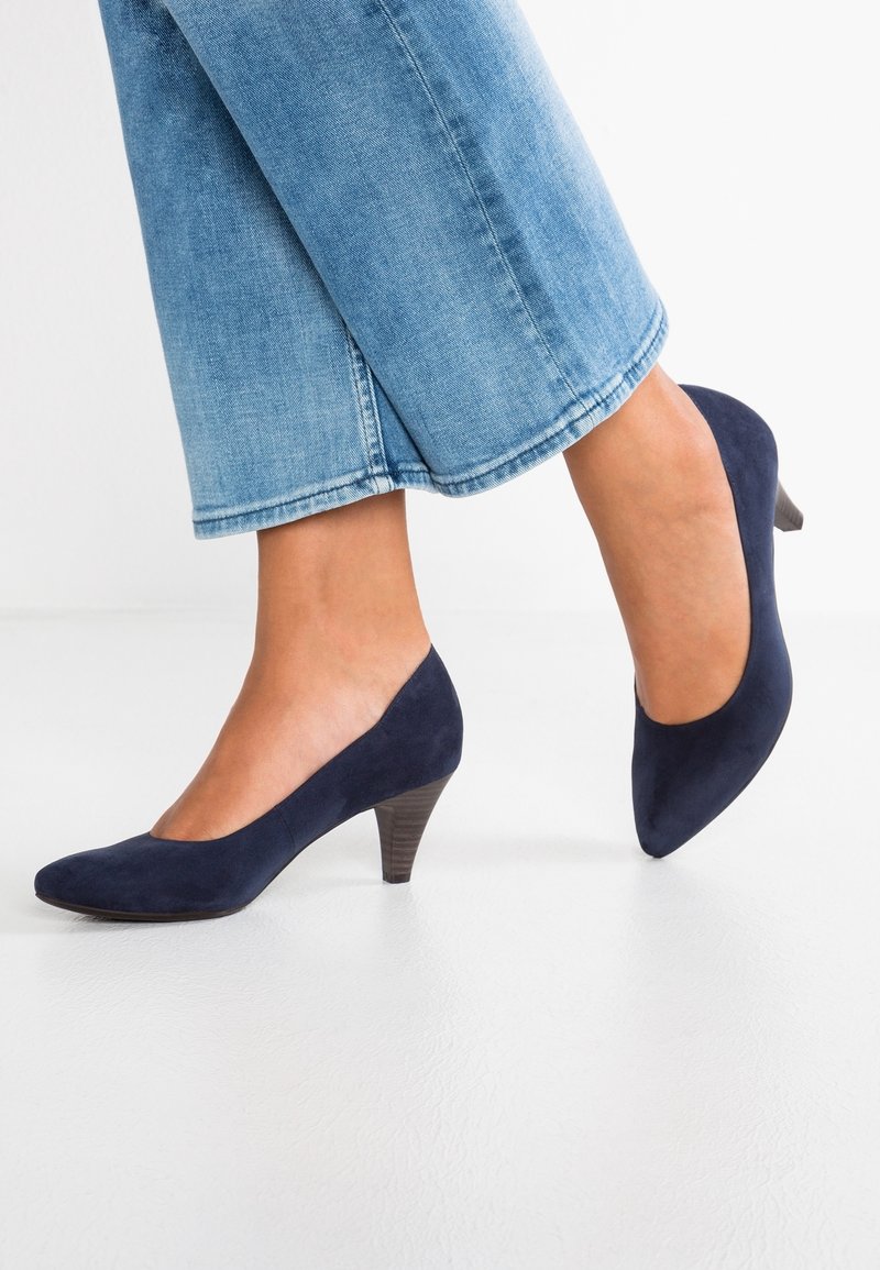Femme portant des chaussures en daim bleu moyen à talon bas, bouts pointus et jean en denim bleu clair raccourci sur un sol blanc.