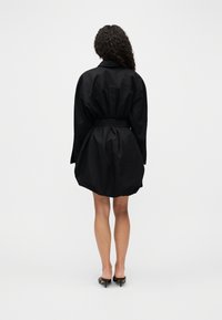 Γυναίκα με σγουρά μαλλιά στέκεται με την πλάτη, φορώντας ένα μαύρο φόρεμα με ζώνη και μαύρα mules με τακούνι, πάνω σε λευκό μονόχρωμο φόντο.