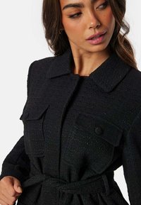 Sorte tekstureret jakke med krave, to forlommer og en livrem for at fremhæve formen. Enkel design med minimale detaljer.