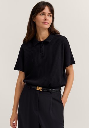 Frau mit schulterlangem braunem Haar, die ein schwarzes, kurzärmeliges, strukturiertes Poloshirt und schwarze Hosen mit einem goldenen Gürtel trägt.