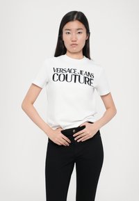 Λευκό κοντομάνικο βαμβακερό t-shirt με μεγάλο μαύρο εκτύπωτο λογότυπο "VERSACE JEANS COUTURE" στο μπροστινό μέρος, συνδυασμένο με μαύρο παντελόνι.