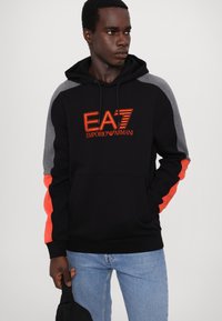 Felpa con cappuccio nera, pannelli grigi sulle spalle e dettagli arancioni. Presenta il logo EA7 arancione, tasca frontale e cappuccio regolabile con cordino.