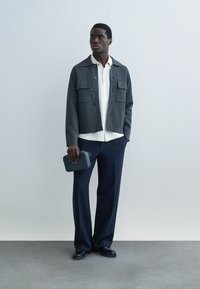 Mann trägt eine dunkelgraue, geknöpfte Jacke, weißes Hemd, marineblaue Hose, schwarze Schuhe, hält eine kleine dunkle Tasche und steht vor einer schlichten hellen Wand.