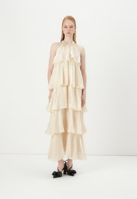 Light cream-colored, tiered maxi dress with a halter neck design. Satin-like texture, flowing layers, and black heeled shoes with bow accents.