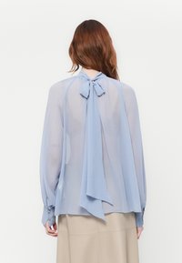 Vrouw met lang bruin haar, gekleed in een doorschijnende lichtblauwe blouse die met een strik rond de nek is geknoopt, gecombineerd met een beige rok, die met de rug naar de kijker kijkt.