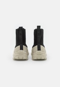 Botas de tobillo de tela negra con suela beige gruesa. Con un tirador en la parte trasera y detalle del logo, resaltado por una textura contrastante.