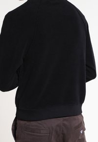 Sudadera negra forrada de lana polar con cuello redondo, mangas raglán y puños y dobladillo de canalé. Textura suave, sin patrones ni adornos visibles.