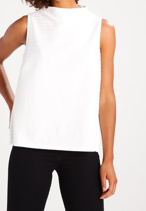 Hvid ærmeløs top med høj hals, med lodrette teksturerede striber. Bæres med sorte bukser, der har en afslappet pasform.