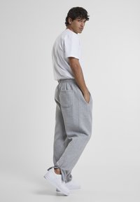 Junger Mann trägt ein weißes T-Shirt, graue Sweathosen mit Gesäßtasche und Reißverschlüssen an den Knöcheln sowie weiße Sneakers, steht mit den Händen in den Taschen.