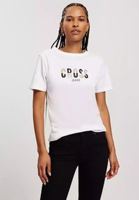 Biała koszulka z krótkim rękawem z nadrukiem "CROSS JEANS" w czarnych i pomarańczowych literach. Połączona z czarnymi dżinsami. Gładki bawełniany materiał.