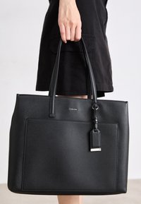 Personne tenant un sac cabas en cuir noir Calvin Klein avec deux poignées et une poche extérieure, portant une jupe ou une robe noire.