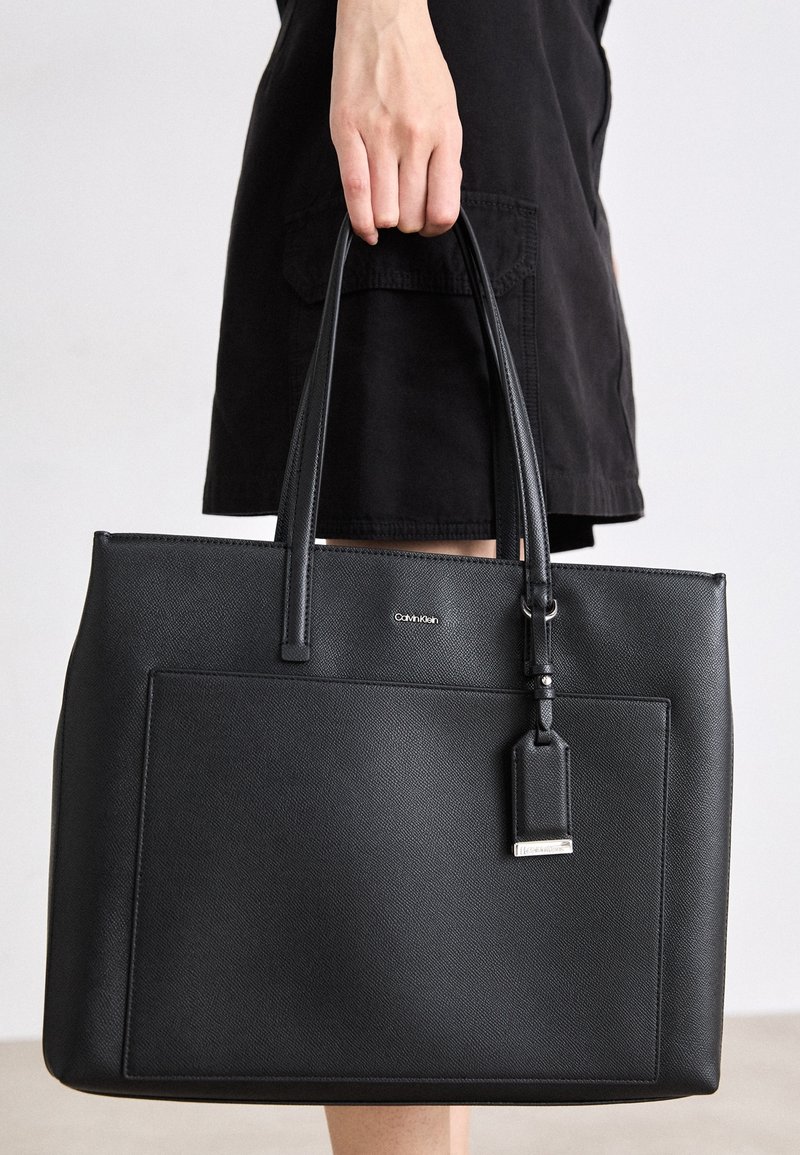 Personne tenant un sac cabas en cuir noir Calvin Klein avec deux poignées et une poche extérieure, portant une jupe ou une robe noire.