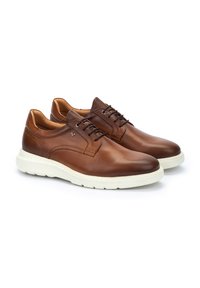 Chaussures en cuir marron avec une finition lisse, semelles en caoutchouc blanches et lacets noirs. Présentent un design minimal et un logo discret sur le côté.