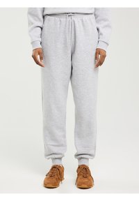 Asmuo dėvi šviesiai pilkas laisvalaikio kelnes su raišteliais ir rudus sportinius batelius, stovintis priešais paprastą šviesią foną.