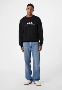 Felpa nera Fila con logo bianco e rosso, abbinata a jeans azzurri a gamba dritta e sneakers bianche con suole color cuoio.