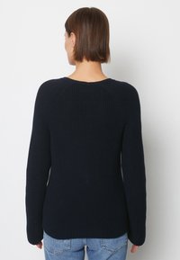 Kvinna med kort brunt hår som bär en svart virkad tröja och ljusblå jeans, sedd bakifrån mot en helt vit bakgrund.