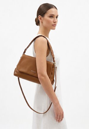 Vrouw in een witte mouwloze jurk draagt een bruine leren schoudertas met gevlochten riem en lange crossbodyriem.