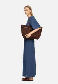 Brązowa zamszowa torba tote z srebrnym okuciem, zamek błyskawiczny i odpinany pasek. Modelka ma na sobie niebieską sukienkę i brązowe baleriny.