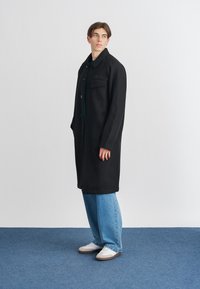 Cappotto di lana nera con colletto, chiusura frontale a bottoni, unica tasca sul petto, maniche a costine, abbinato a jeans celesti e sneakers bianche.