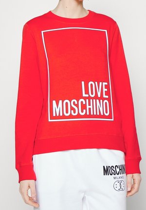 Osoba nosząca czerwoną bluzę z białym napisem „LOVE MOSCHINO” w obrębie obrysu kwadratu, w połączeniu z białymi spodniami z czarnym napisem „MOSCHINO MILANO”.