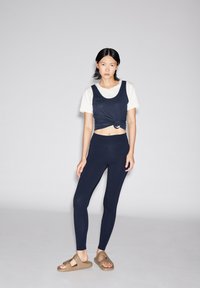 Marineblauw crop tanktop met een geknoopt detail over een wit T-shirt, gecombineerd met marineblauwe leggings en bruine sandalen tegen een lichtgrijze achtergrond.