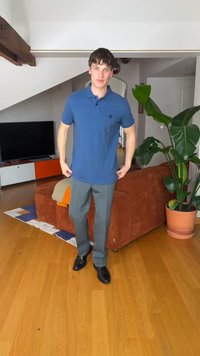 Νέος άνδρας που φορά μπλε πόλο μπλούζα και γκρι παντελόνι στέκεται σε σαλόνι με ξύλινο δάπεδο, πορτοκαλί καναπέ, μεγάλο φυτό και τηλεόραση στο φόντο.