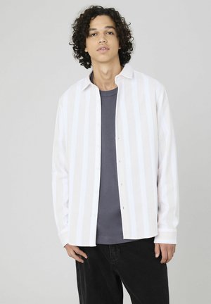 Chemise à rayures blanches avec des boutons, en tissu léger, comprenant un col, des manches longues et une coupe décontractée, portée sur une chemise gris foncé.