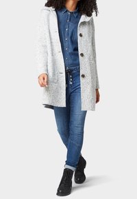 Žena v světle šedém strukturovaném kabátu přes modrou denimovou košili, s džínami a černými šněrovacími botami, stojí se zkříženou jednou nohou vpředu.