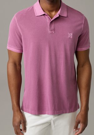 Mann trägt ein kurzärmeliges, mauvefarbenes Poloshirt mit Knöpfen und weiße Hose vor einem schlichten grauen Hintergrund.