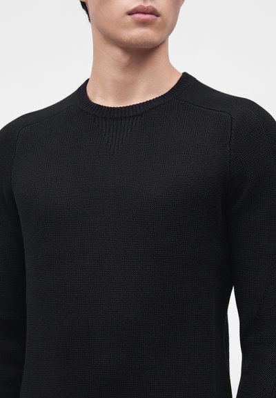 Jeune personne porte un pull en maille noire texturée à col rond, présenté des épaules au menton sur un fond clair uni.