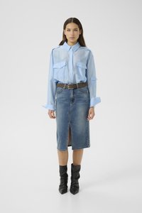 Woman wearing a sheer light blue button-up shirt, belted denim skirt with front slit, and black pointed-toe ankle boots, standing against plain background.