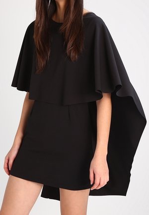 Femme portant une mini robe noire avec un drapé style cape s'étendant des épaules jusqu'à mi-cuisse, debout devant un fond uni.