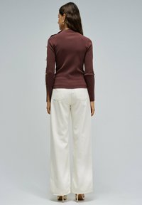 Suéter marrón ajustado con cuello alto y detalles de botones en las mangas, combinado con pantalones anchos blancos. Zapatos planos visibles.