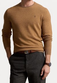 Senapsgul stickad tröja med rund halsringning och långa ärmar, med en liten logga på bröstet. Bärs ihop med gråa byxor och ett brunt bälte.