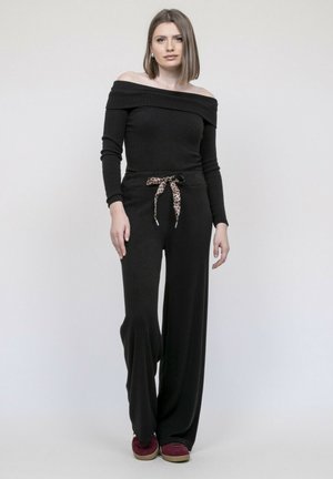 Jeune femme en haut noir à épaules dénudées et pantalon large avec un foulard imprimé léopard, posant devant un fond clair uni.