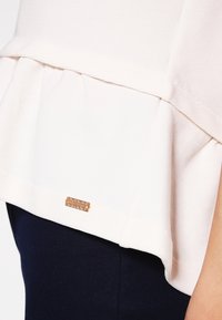 Ljusrosa topp med volangkant, tillverkad av strukturerad tyg. Har en liten metallbricka märkt "ROSS + BRACE" nära den nedre sömmen.