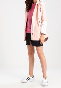Frau, die einen rosa gestrickten Pullover, eine hellrosa Regenjacke, einen schwarzen Jeansrock mit Schlitz vorne und weiße Adidas-Sneaker mit schwarzen Streifen trägt.