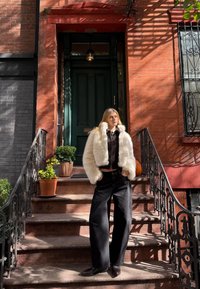 Bijela umjetna krznena jakna, crne široke traperice, crne cipele. Crvene ciglene stepenice, zelena vrata s dekorativnim crnim ogradom, saksije s biljkama.