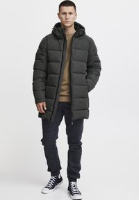 Tamnozelena puf jakna s kapuljačom, kockasti dizajn, prednji zip, i bočne džepove, nosi se preko smešnog dukserice i crnih pantalona.