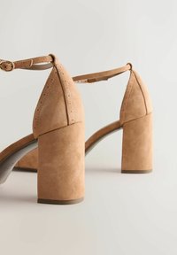Beige Wildleder-Sandalen mit hohem, klobigem Blockabsatz, Knöchelriemen und spitzer Zehenpartie mit perforierten Details am Obermaterial.