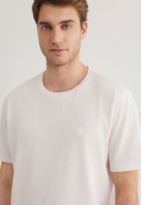 Light pink short-sleeve t-shirt made from soft fabric. Features a rounded neckline and a white logo on the left chest area.