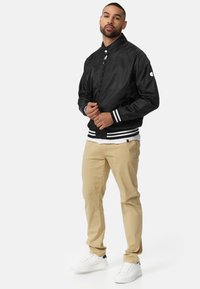 Chaqueta bomber negra con puños y dobladillo blanco a rayas, usada sobre una camisa blanca, combinada con pantalones caqui y zapatillas blancas.