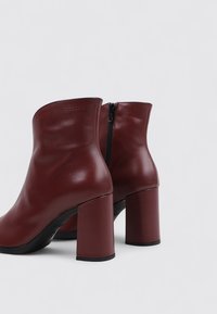 Botas de tobillo de cuero burdeos con una textura suave, que cuentan con un cierre de cremallera y tacones cuadrados gruesos. Líneas limpias y diseño minimalista.