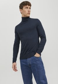 Pulover tricotat cu guler înalt, de culoare bleu marin, cu detalii cu nervuri la guler și la margine, asortat cu blugi de denim de culoare bleu deschis.