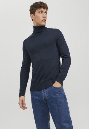 Suéter de punto color azul marino con cuello de tortuga y detalles acanalados en el cuello y el dobladillo, combinado con jeans de mezclilla de color azul claro.