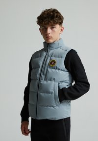 Gilet matelassé bleu clair avec col montant et fermeture éclair, arborant un patch logo rond, associé à un t-shirt à manches longues noir.